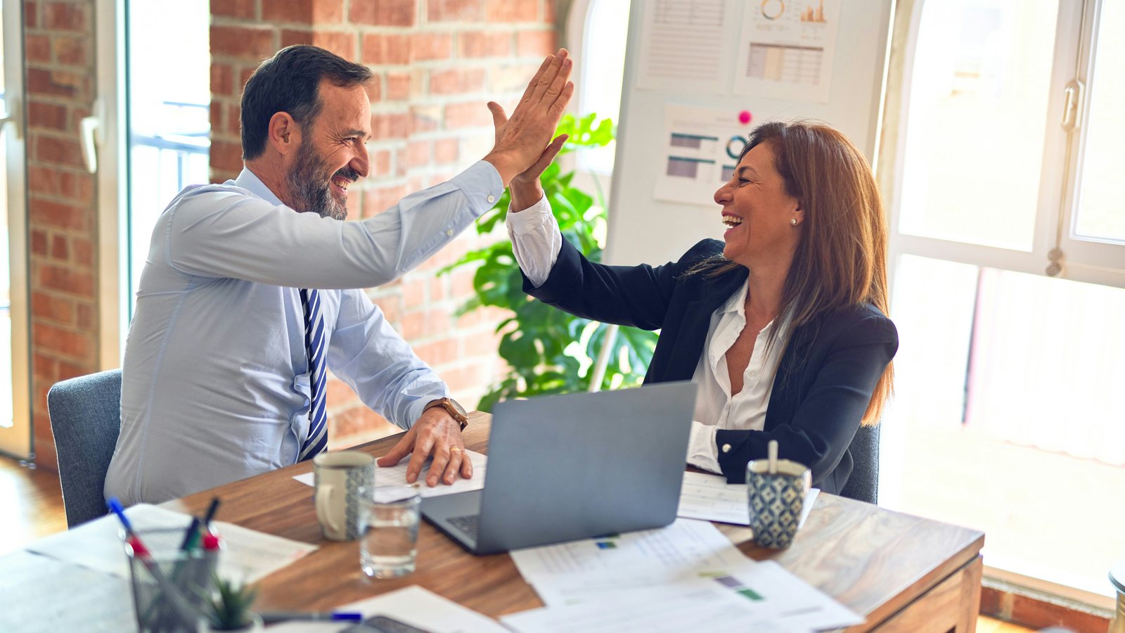 Twee collega's geven elkaar een high five tijdens een overleg met laptop en documenten
